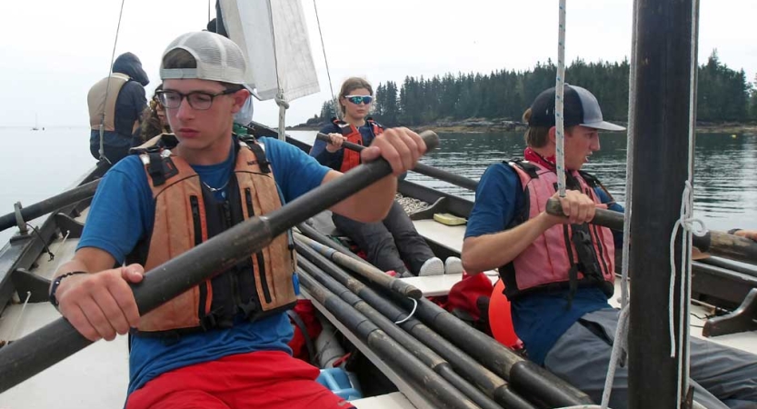 Teens sitting in a sailboat use oars. They are all wearing life jackets. 